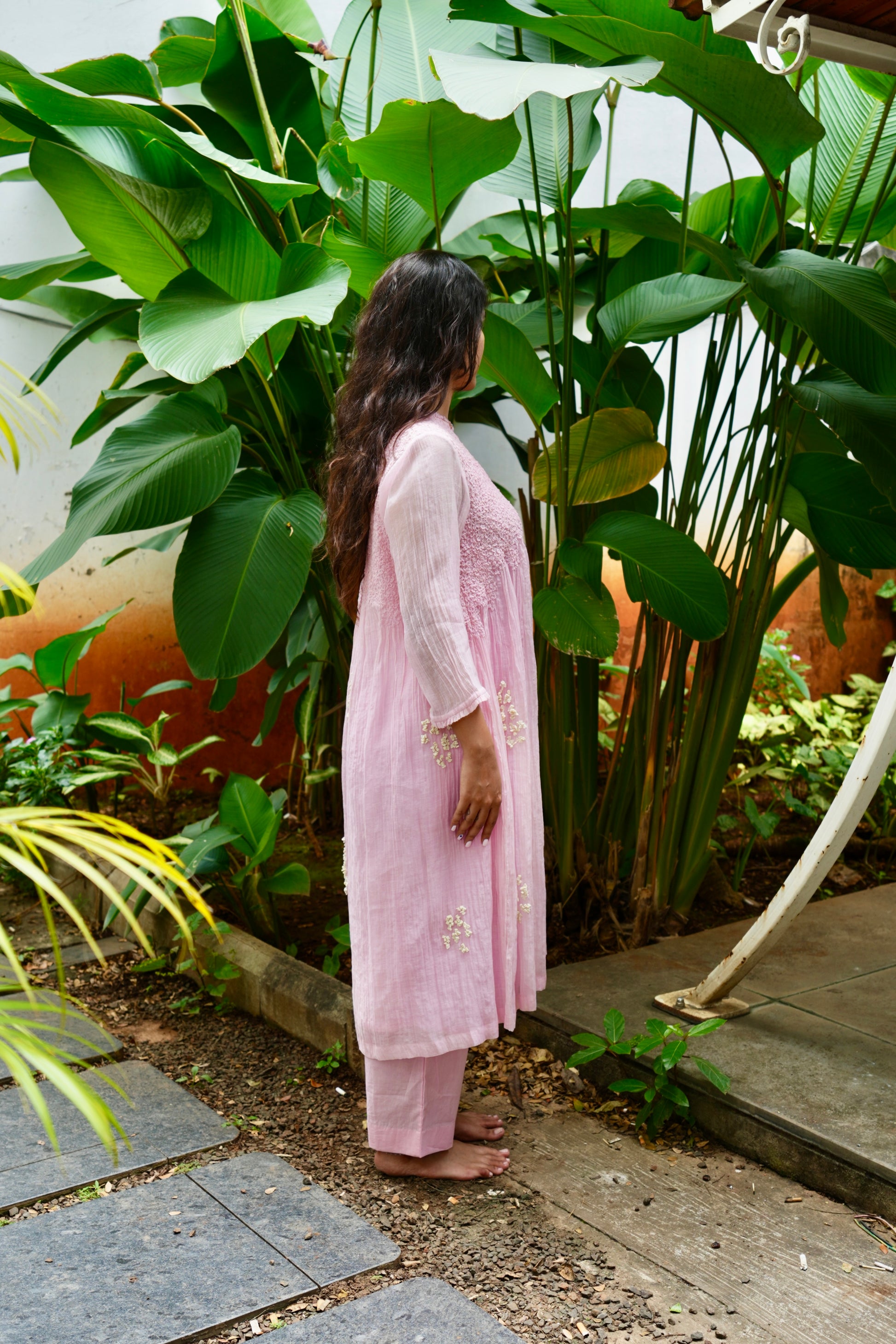 Woman in a pink traditional outfit standing in a garden with large green leaves.