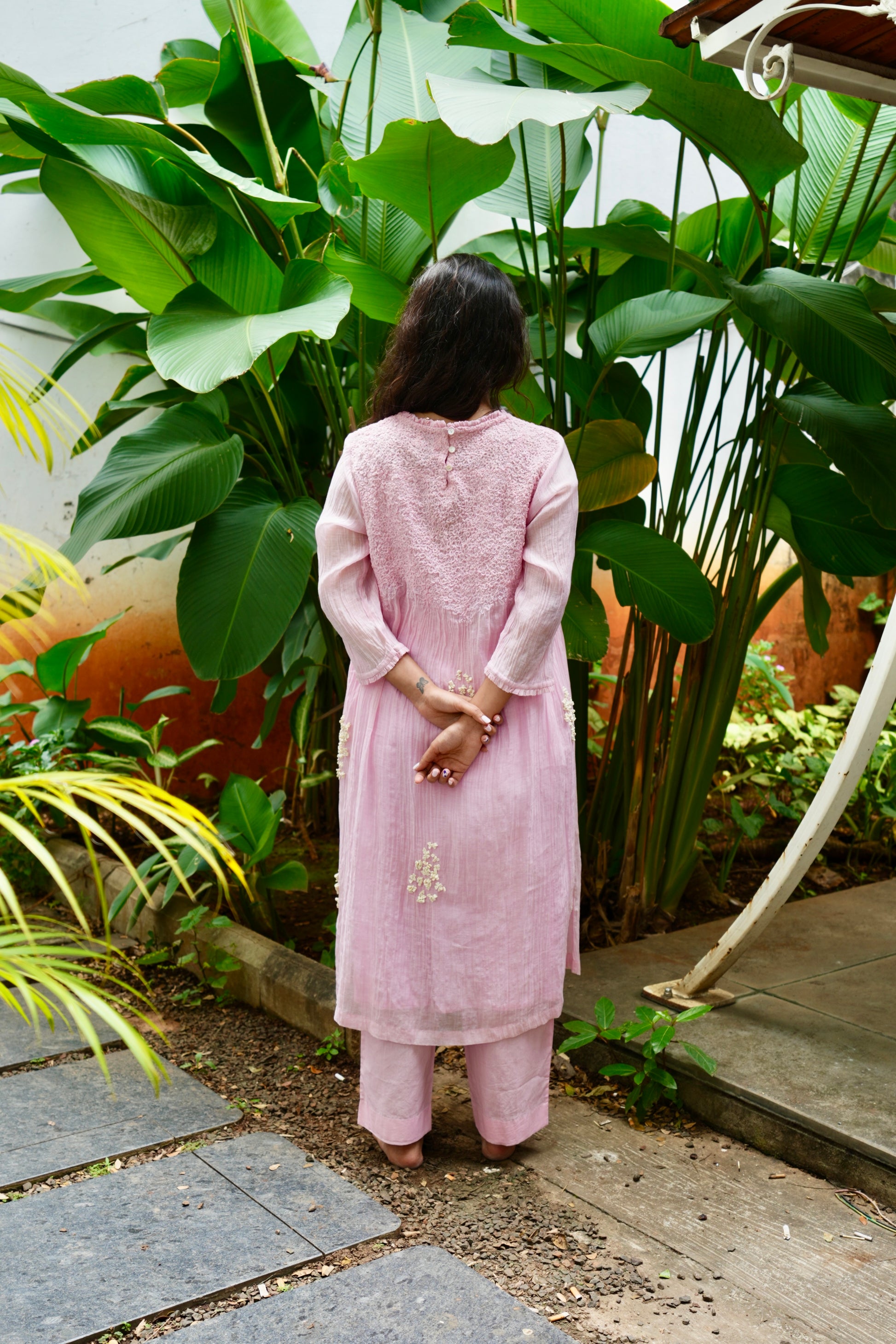 Person wearing a pink traditional outfit standing in a garden with green plants.
