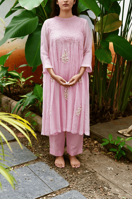 Person wearing a pink dress with floral embroidery standing in a garden setting.